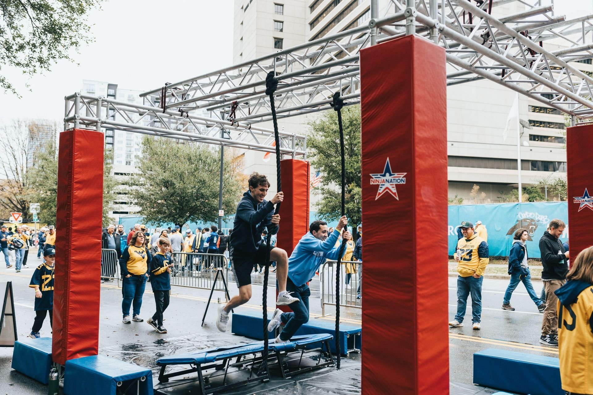 People on an obstacle course. Two people use poles to swing between platforms at an outdoor event.