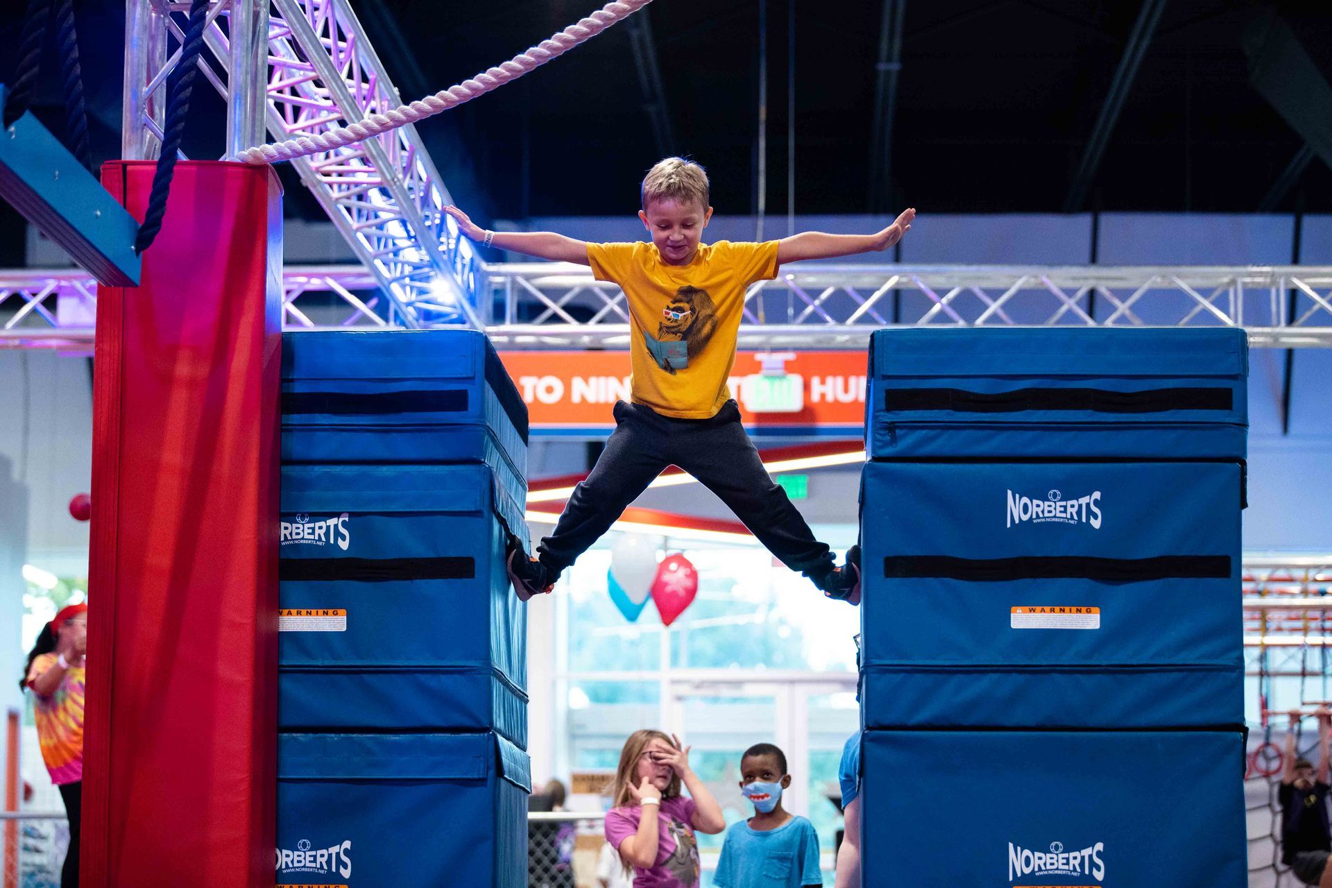 Boy jumping between blue foam blocks at an indoor obstacle course; other children watch.
