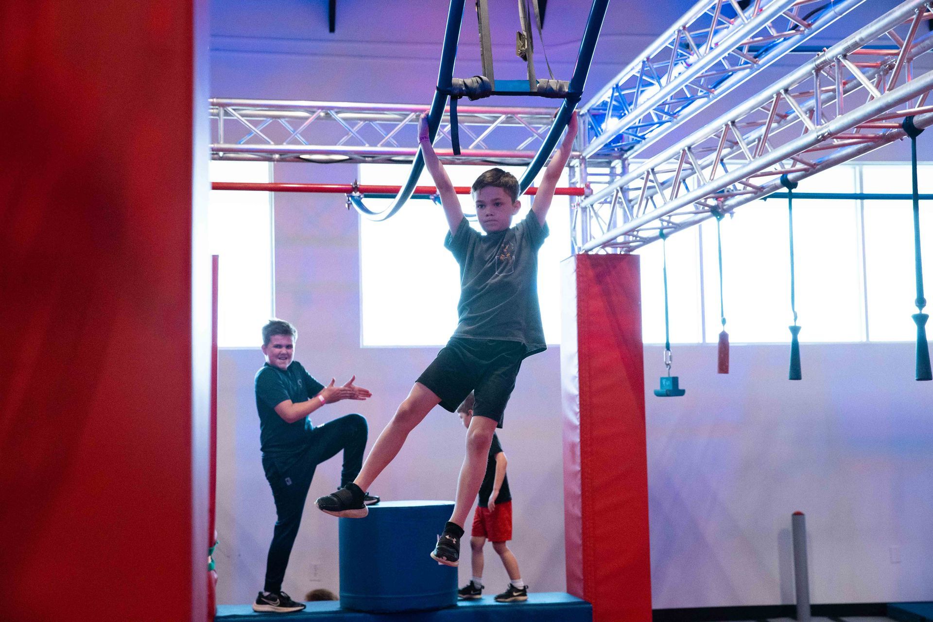 Boy on ninja obstacle, grasping bars, other boy gesturing, indoor gym setting.