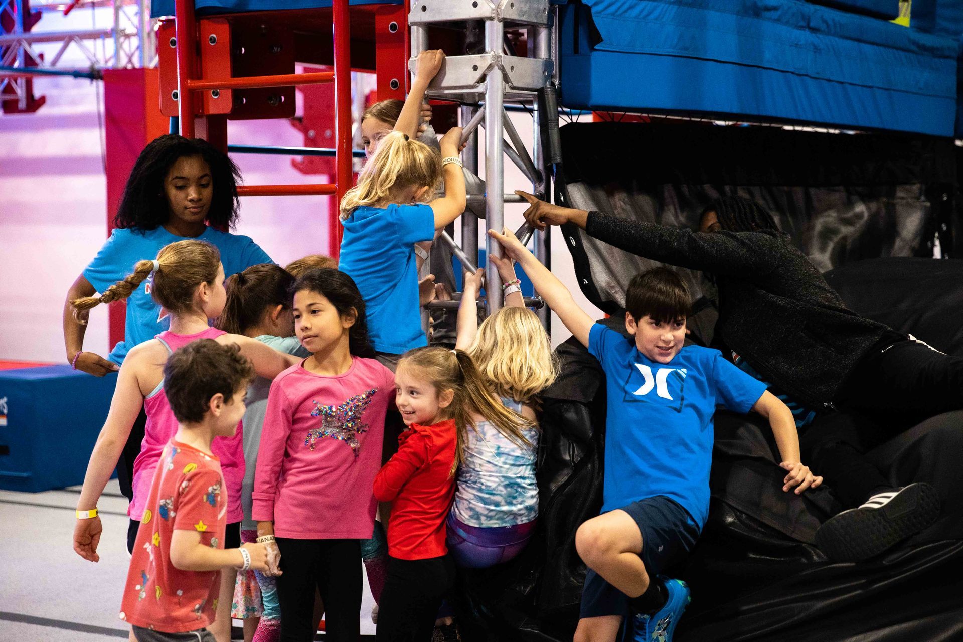 Children with a coach at an indoor obstacle course, reaching for a climbing structure.