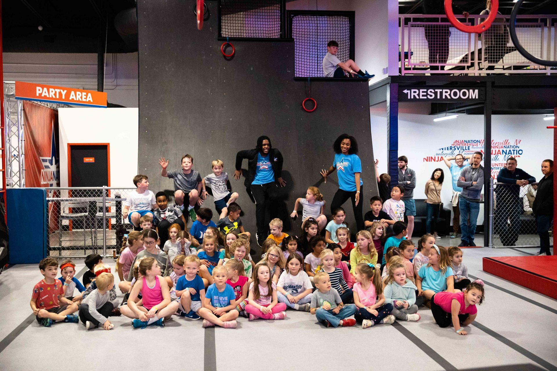 Group of children and adults pose in an indoor ninja warrior gym, smiling, some waving.