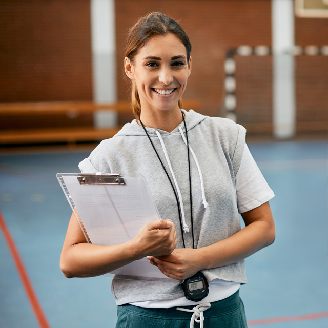 Coach in gym smiling, holding clipboard and stopwatch.