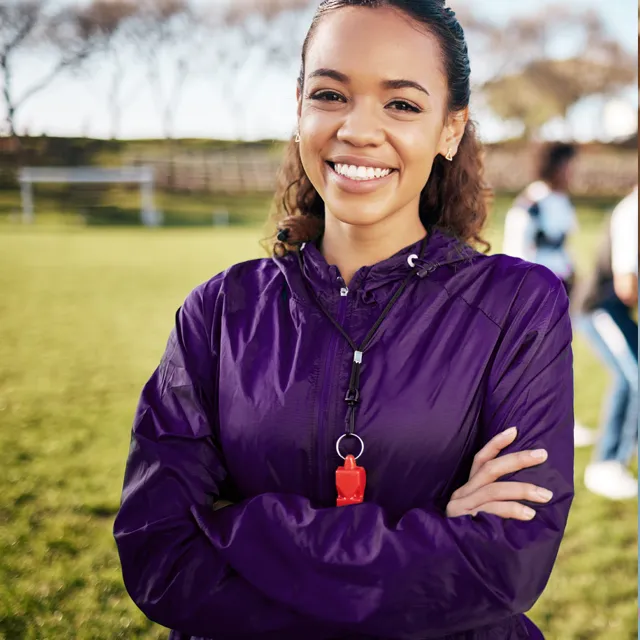 Woman smiling with arms crossed, wearing purple jacket and whistle on a field.