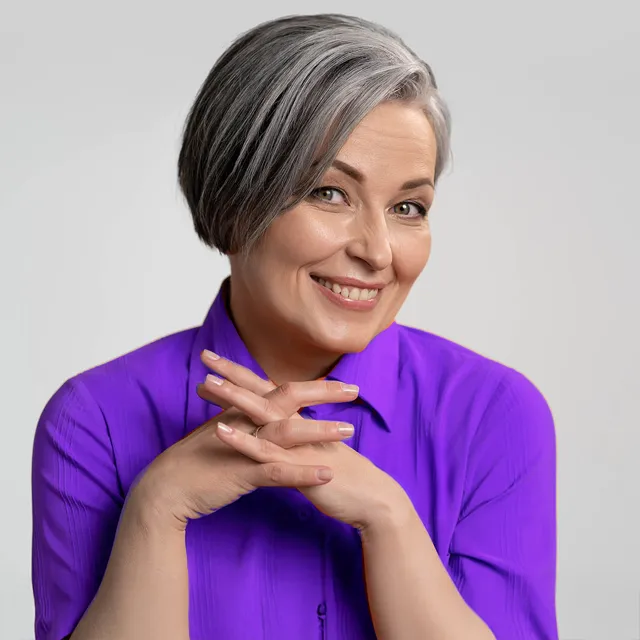Woman with gray hair smiles, wearing a purple shirt, hands clasped.