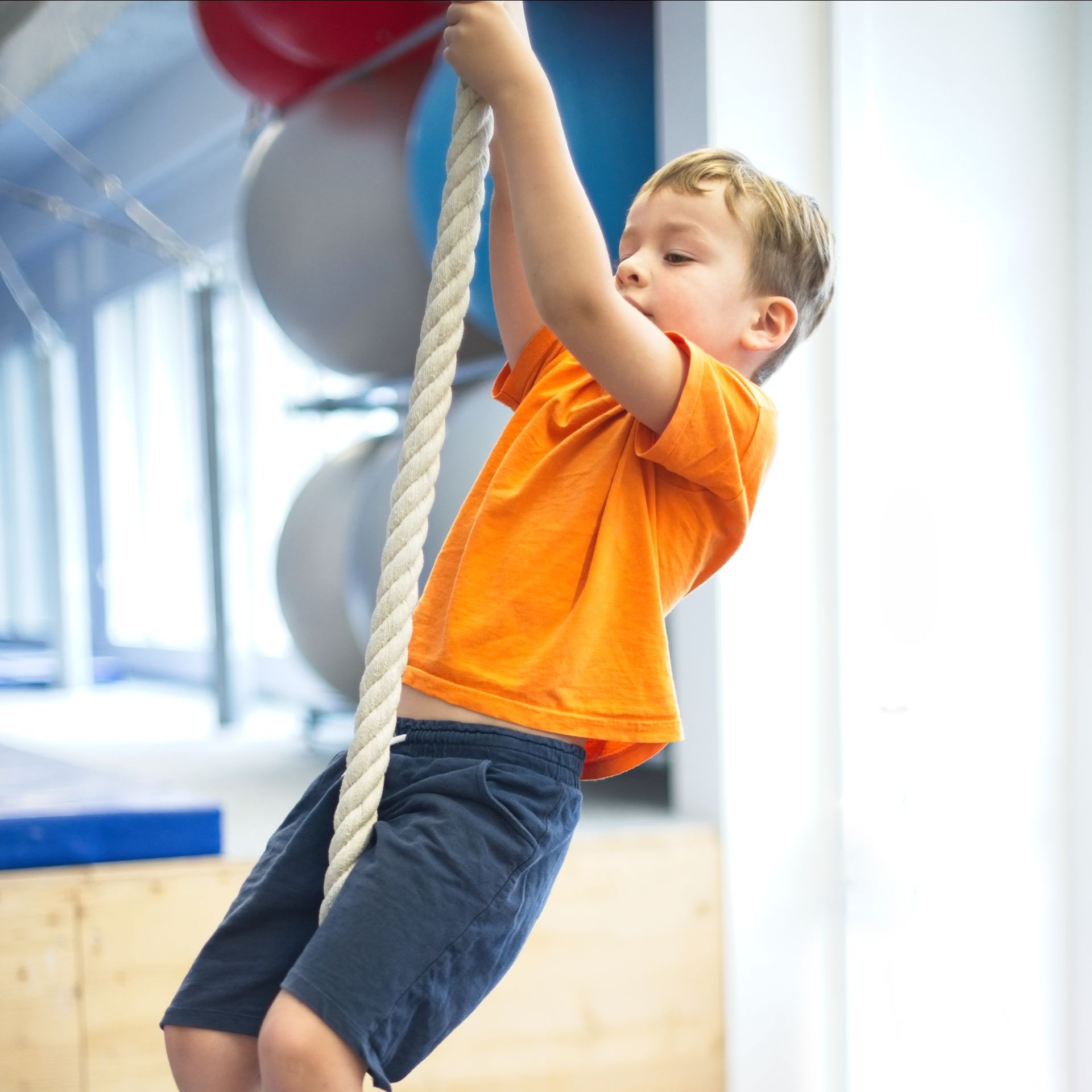 Boy in orange shirt climbs a rope in a gym, holding on with both hands.