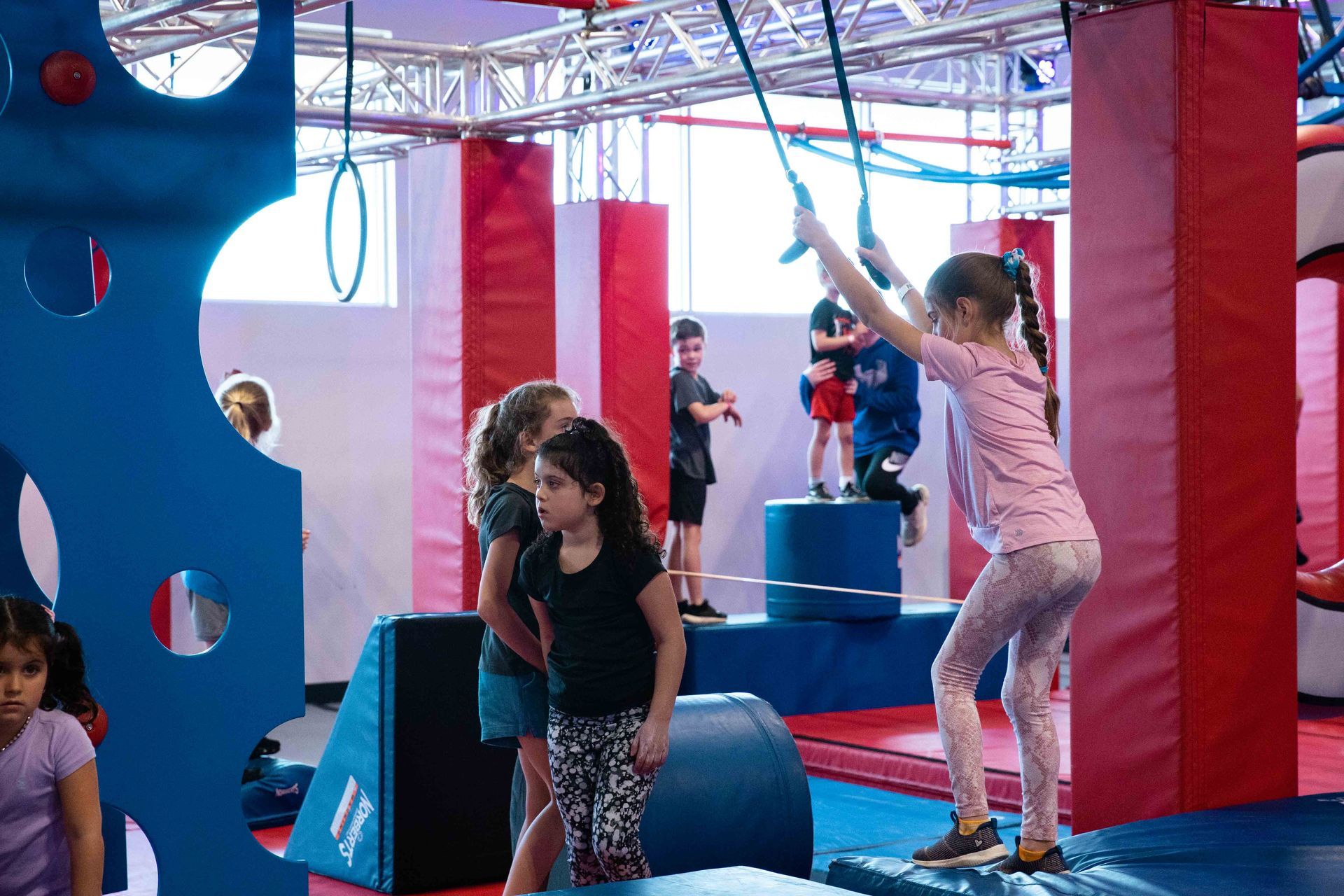 Children play on an indoor obstacle course featuring red padded pillars, blue foam mats, and hanging gymnastic rings.