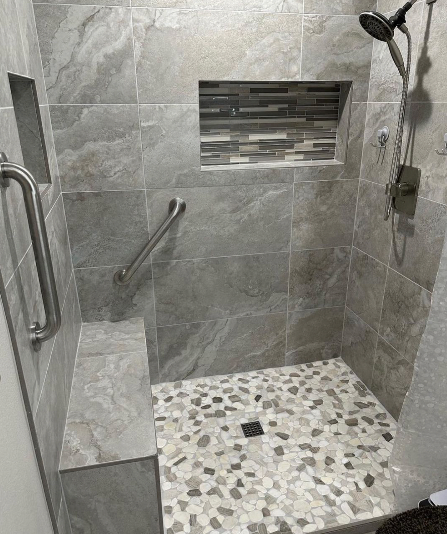 A walk in shower with a stainless steel handle and a tiled floor.