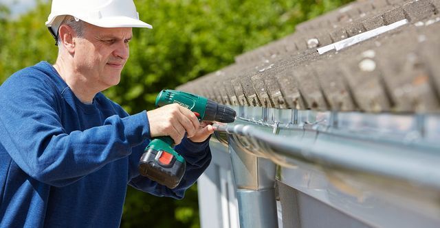 Man in hard hat using a drill to install gutter on a house.
