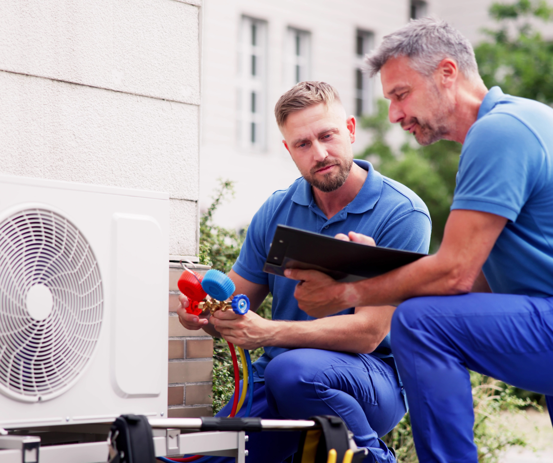 Two technicians in blue uniforms examine an AC unit, one holding a gauge.