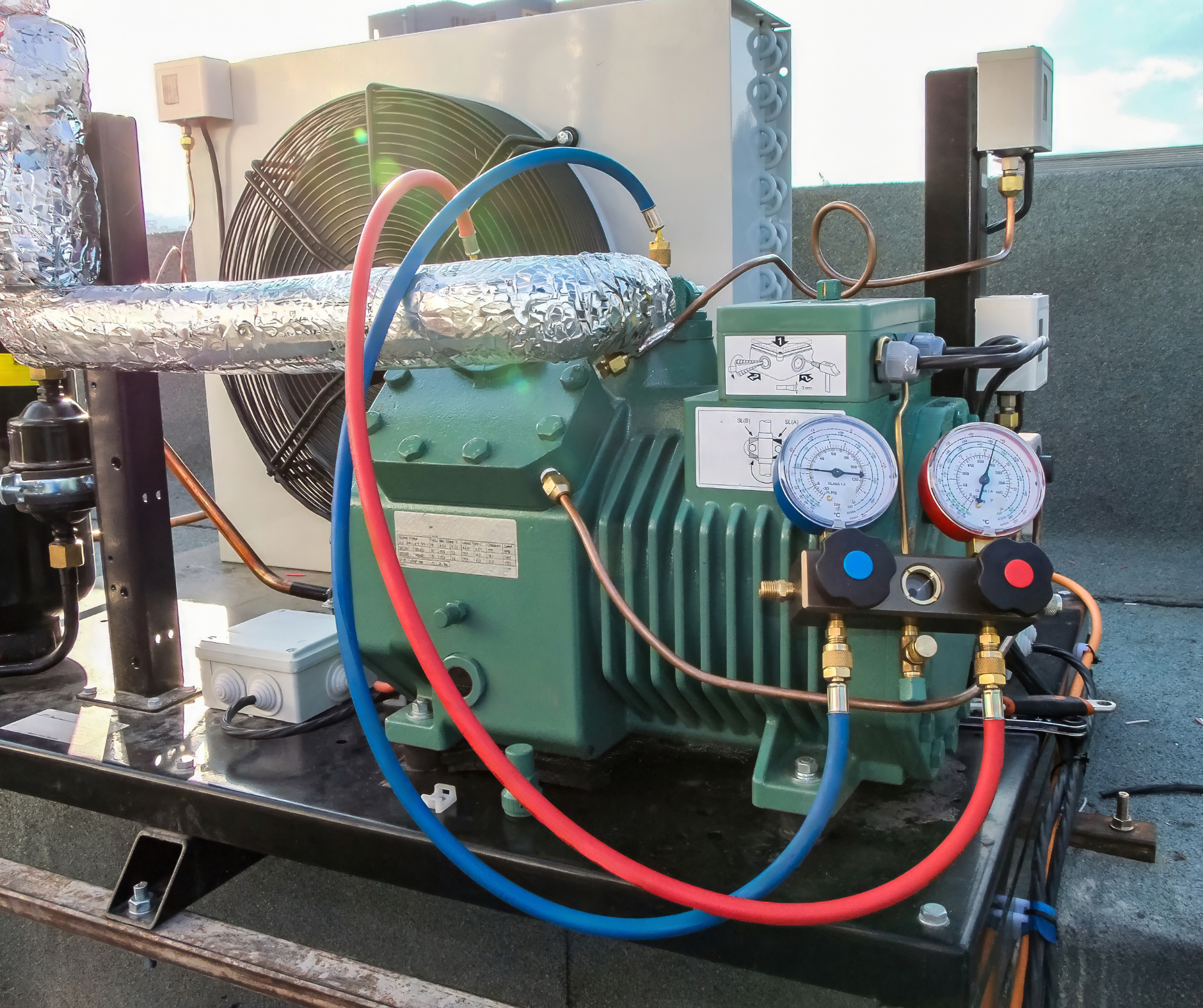 Green refrigeration compressor with gauges and hoses on a rooftop, with a fan and insulated pipe.