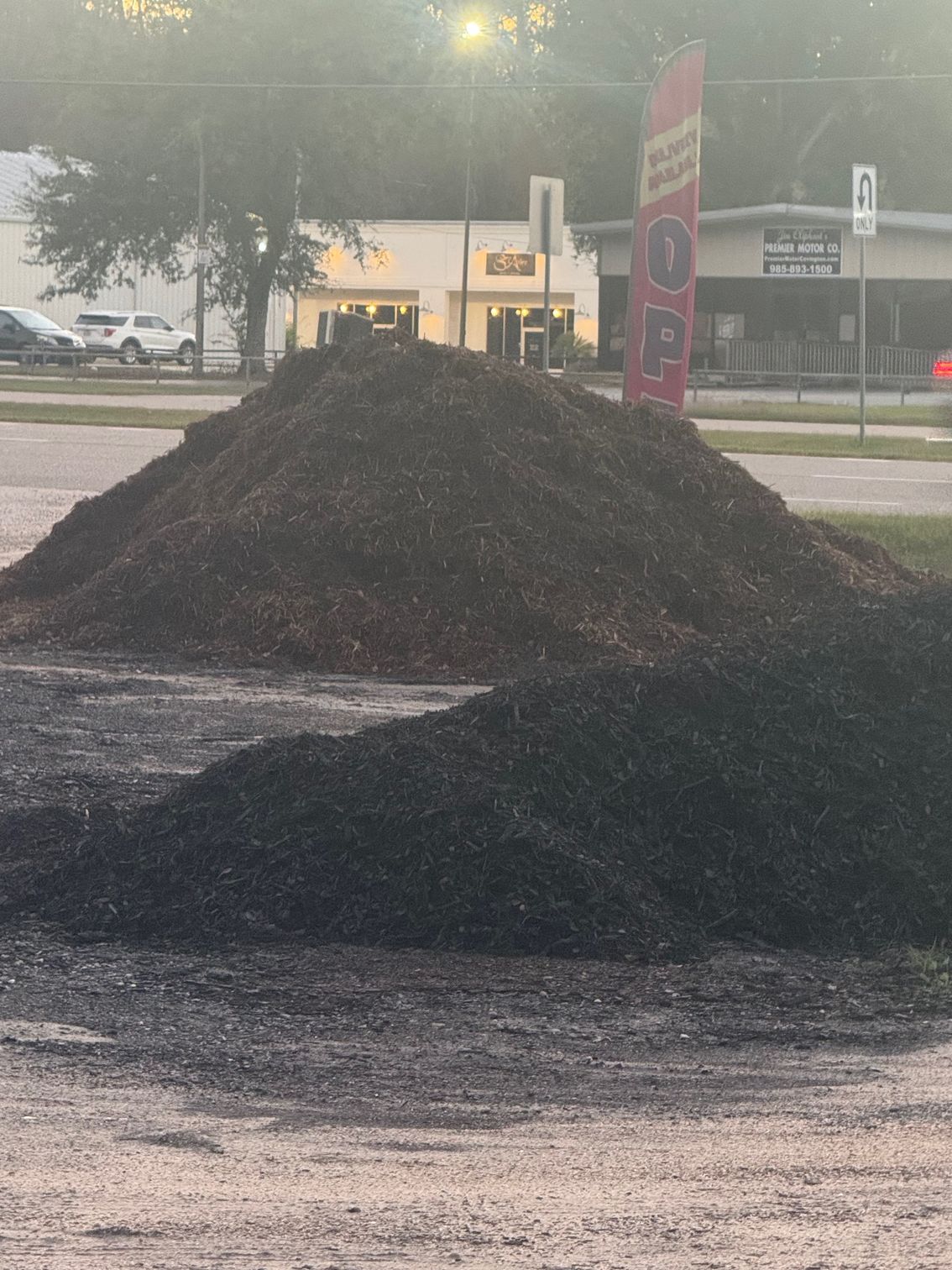 Two large piles of dark mulch sit outside a business. Buildings and a sign are in the background.