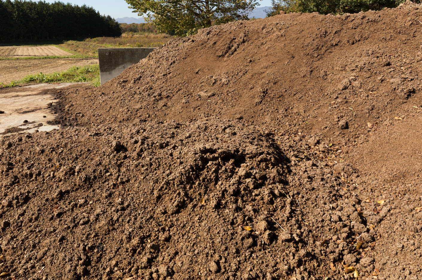 Pile of brown soil or compost outdoors with a field and trees in the background.