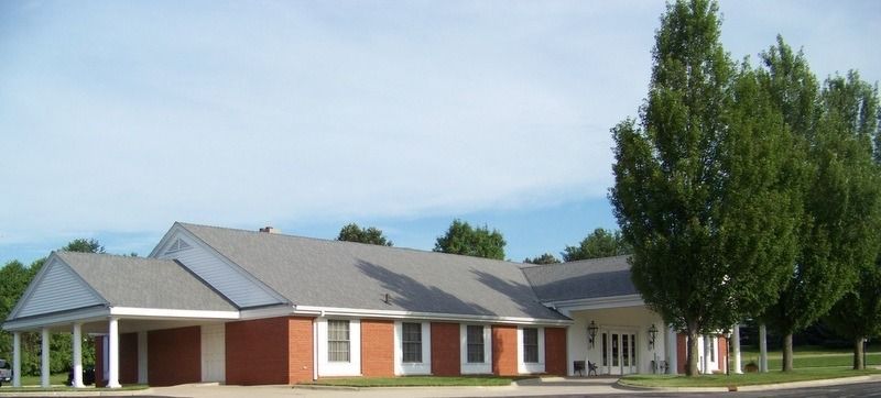 A one-story brick building with a gray roof and tall green trees in front.