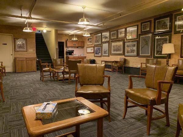 Lounge area with brown leather chairs, framed pictures, and a small bar. Carpeted floor, exposed ceiling.