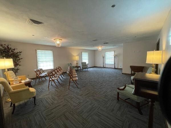Empty room, possibly a funeral home, with chairs, windows, and lamps. Carpeted floor, neutral colors.