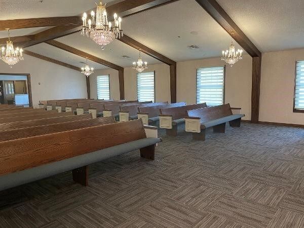 Empty chapel with wooden pews, chandeliers, and exposed beams. Beige walls, carpet, and window blinds.
