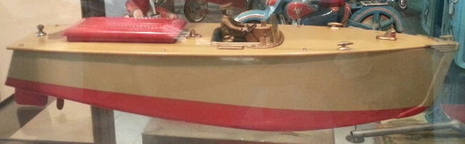 A tan and red model motorboat sits on display, featuring a red seat and a steering wheel.