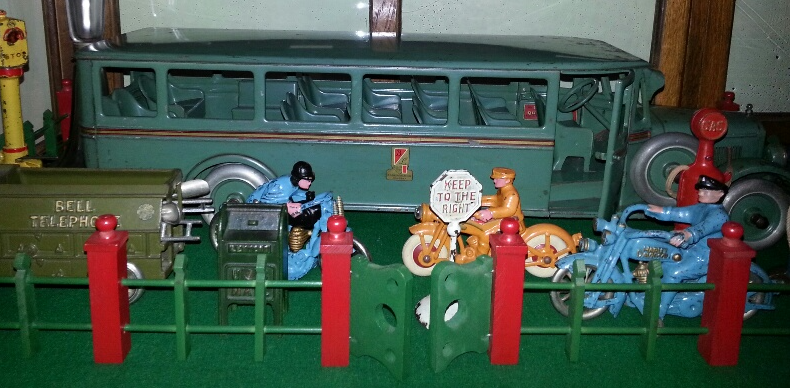 A green toy bus, a trailer, and blue toy motorcycles with riders behind a green and red toy fence.
