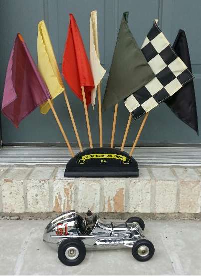 A vintage toy race car in front of a stand holding seven colorful racing flags on a concrete step.