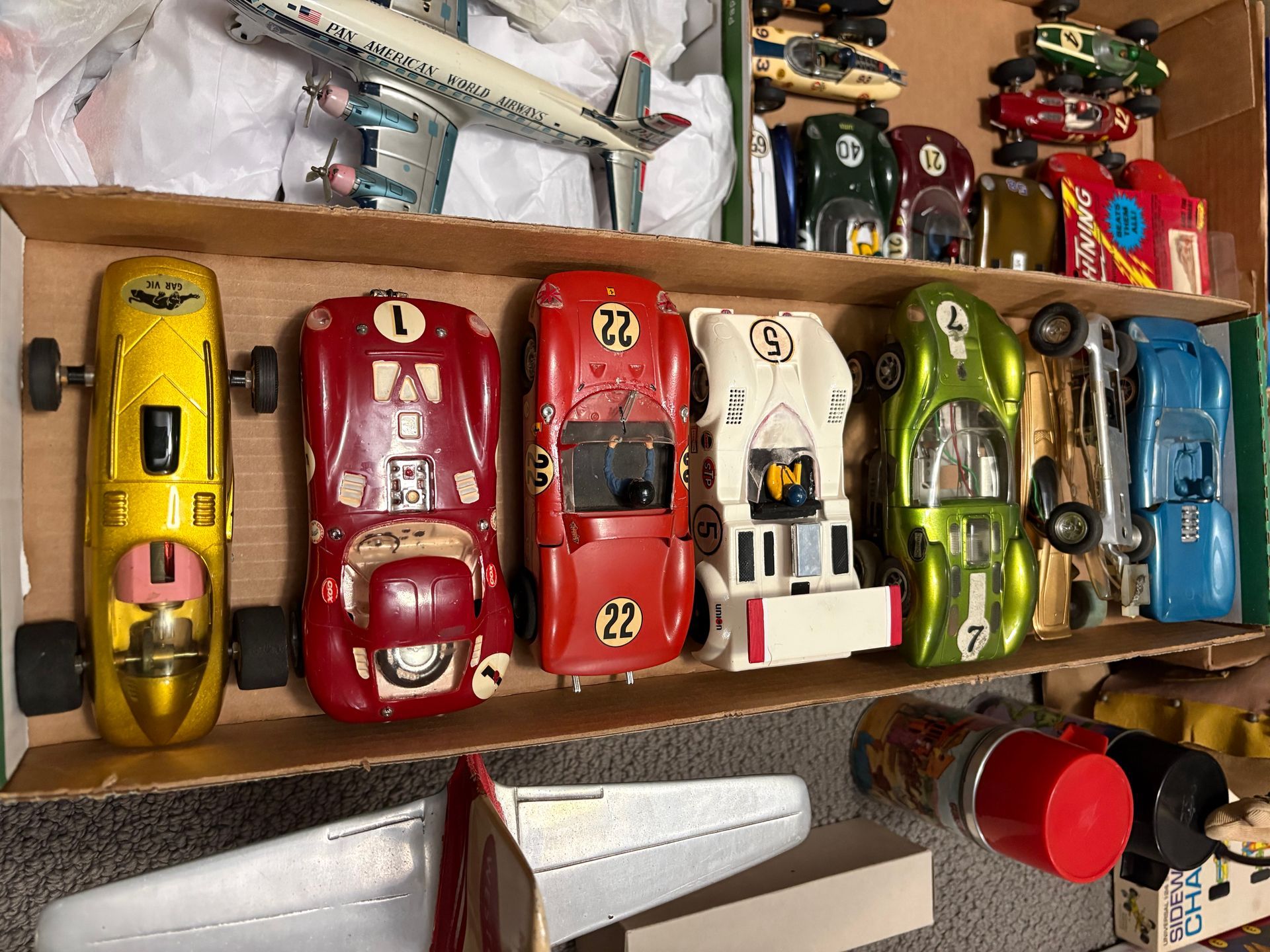 Colorful toy race cars lined up on a shelf, with more toys and a red container nearby.