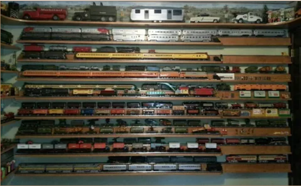A wall-mounted display of numerous model trains and toy vehicles arranged on horizontal wooden shelves.