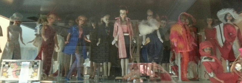 A row of vintage-style dolls wearing diverse fashionable outfits displayed inside a glass cabinet.
