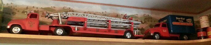 Two toy trucks, a red fire ladder truck and a red and blue delivery truck, sit on a wooden shelf.