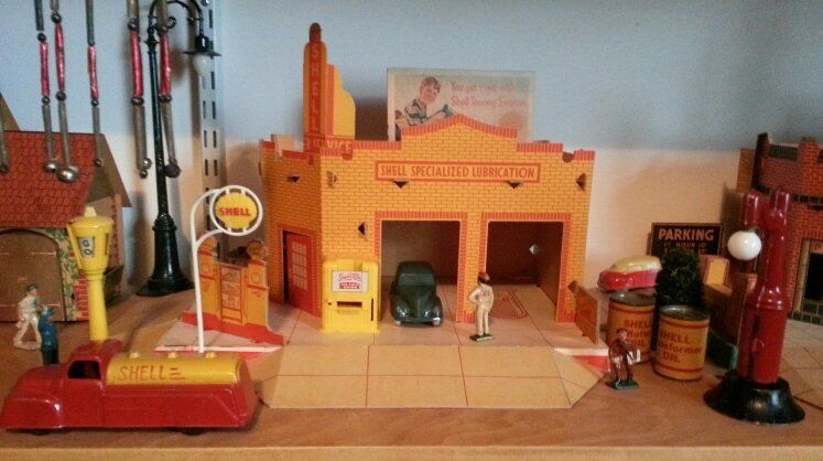 A vintage toy gas station display with a red tanker truck, a parked toy car, miniature figures, and decorative lamps.
