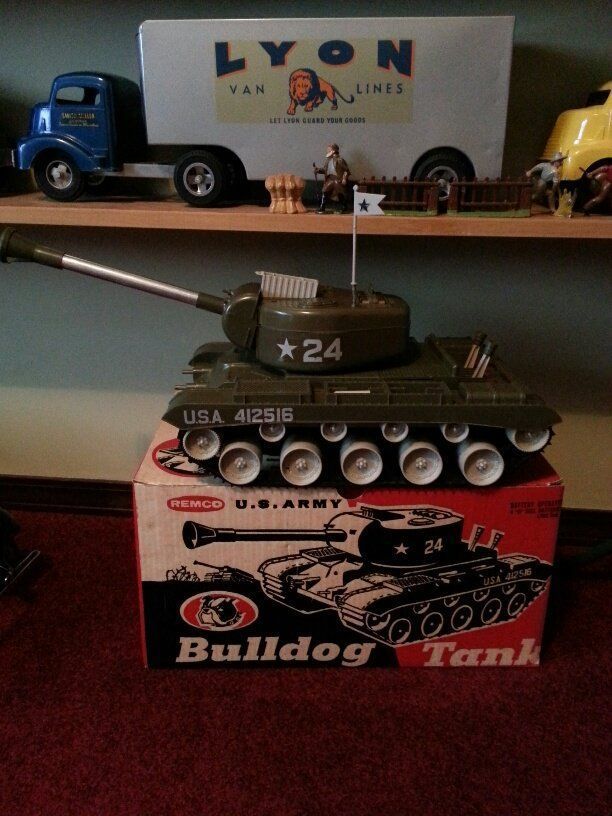 A Renco Bulldog U.S. Army toy tank with a white flag rests on its box, with a Lyon Van Lines toy truck on a shelf behind.