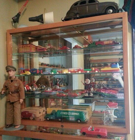 A glass display cabinet filled with vintage toy cars, trucks, and a small figure, topped with a large brown toy car.
