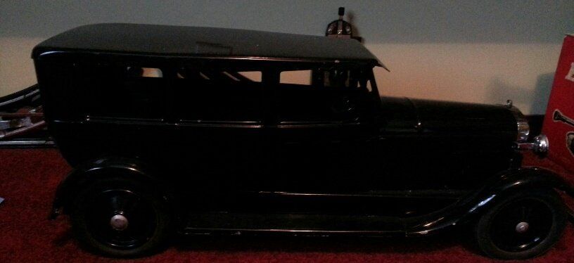 A black antique toy car, viewed from the side, sits on a dark red surface.