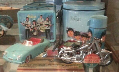 A vintage-style blue toy car and motorcycle displayed near Beatles-themed lunch boxes and a canister on a shelf.