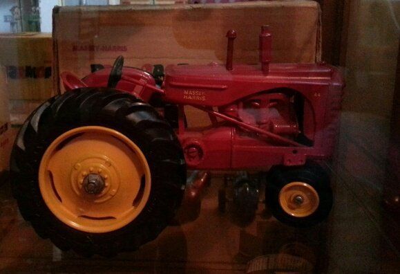A red Farmall toy tractor with large yellow wheels, displayed on a shelf in front of a cardboard box.
