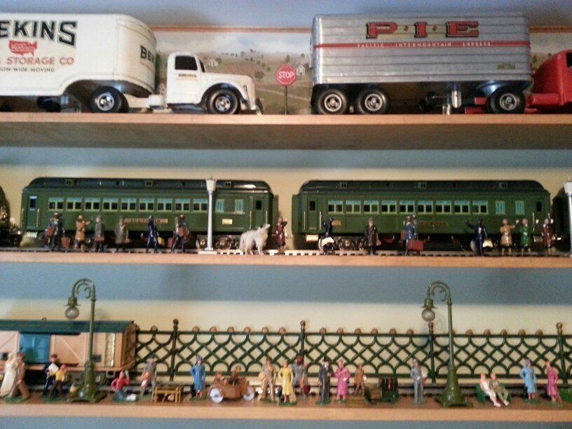 Three wooden shelves display model toy semi-trucks, passenger train cars, and numerous small figurines of people.