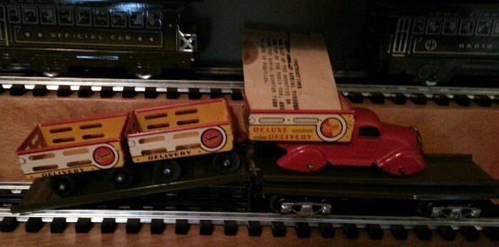 A vintage red toy truck and two yellow livestock trailers are loaded onto a green model train flatcar.