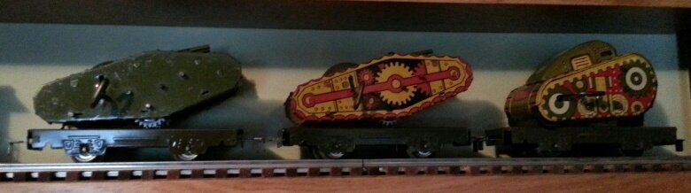 Three vintage toy tanks in different colors sitting on dark metal model train flatbed cars on a shelf.