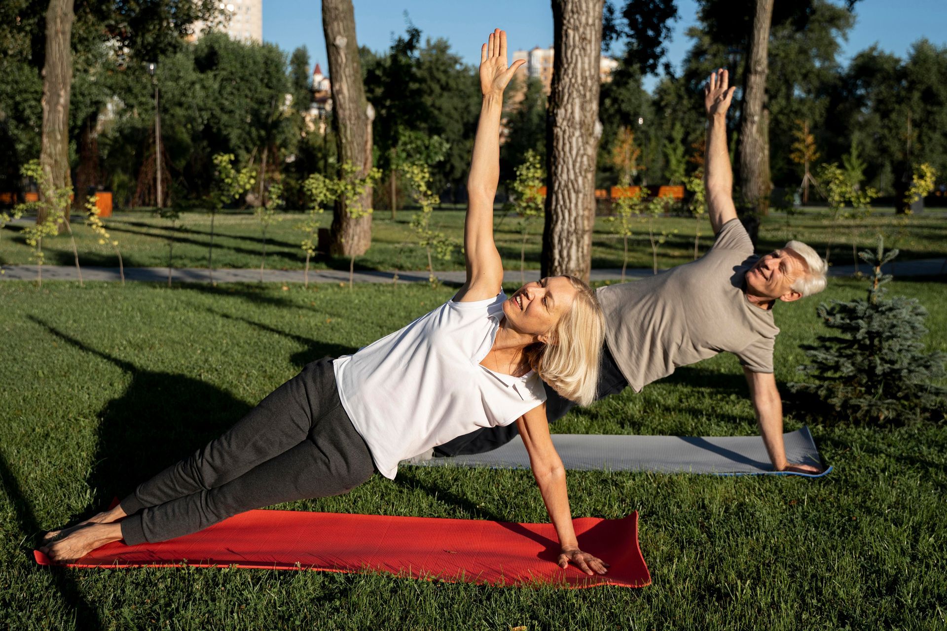 Two people doing side plank yoga pose in a grassy park, arms raised.