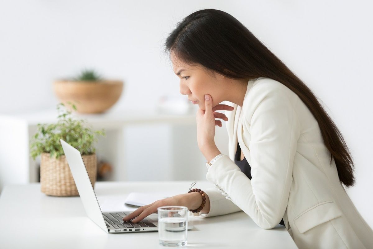 Woman in white blazer, focused on laptop, hand on chin, water glass on desk.