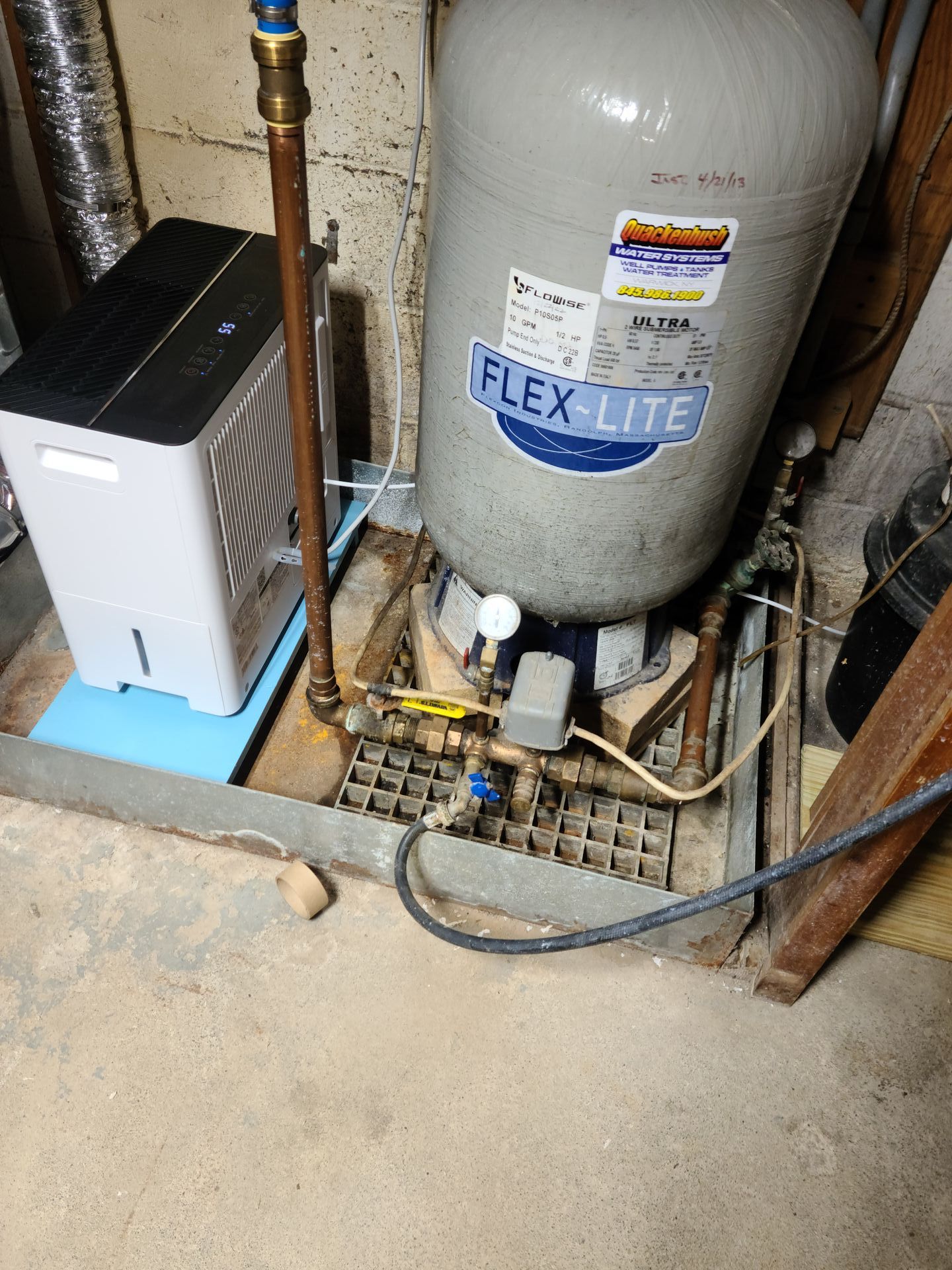 A water pressure tank with attached pipes and a dehumidifier sit in a basement. The tank is gray, and the dehumidifier is white and black.