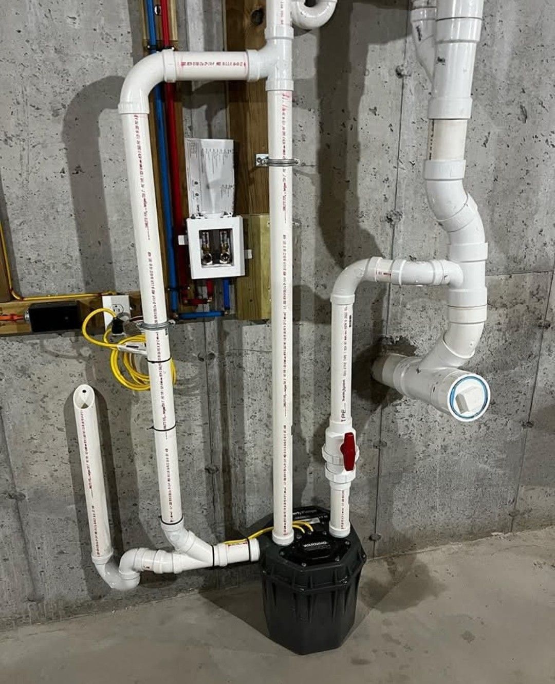 Basement plumbing setup with white PVC pipes, a sump pump, and electrical components against a concrete wall.
