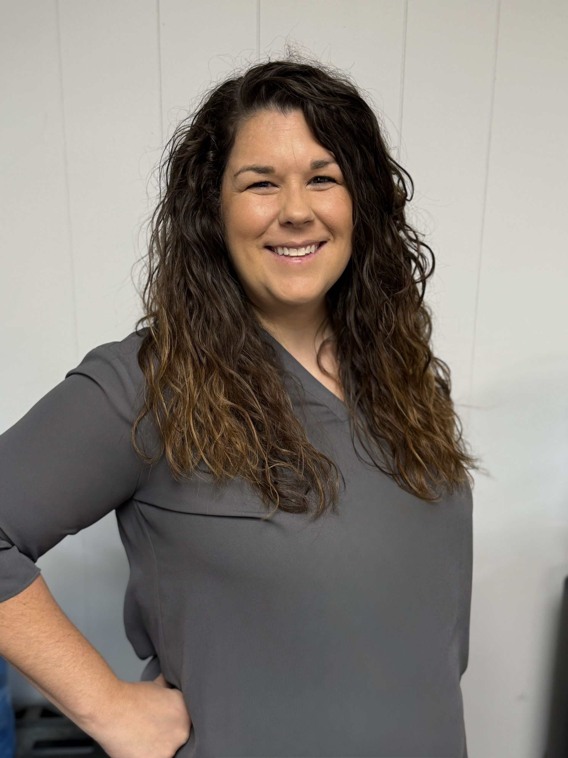 Woman in gray top smiles with hand on hip against a white wall.