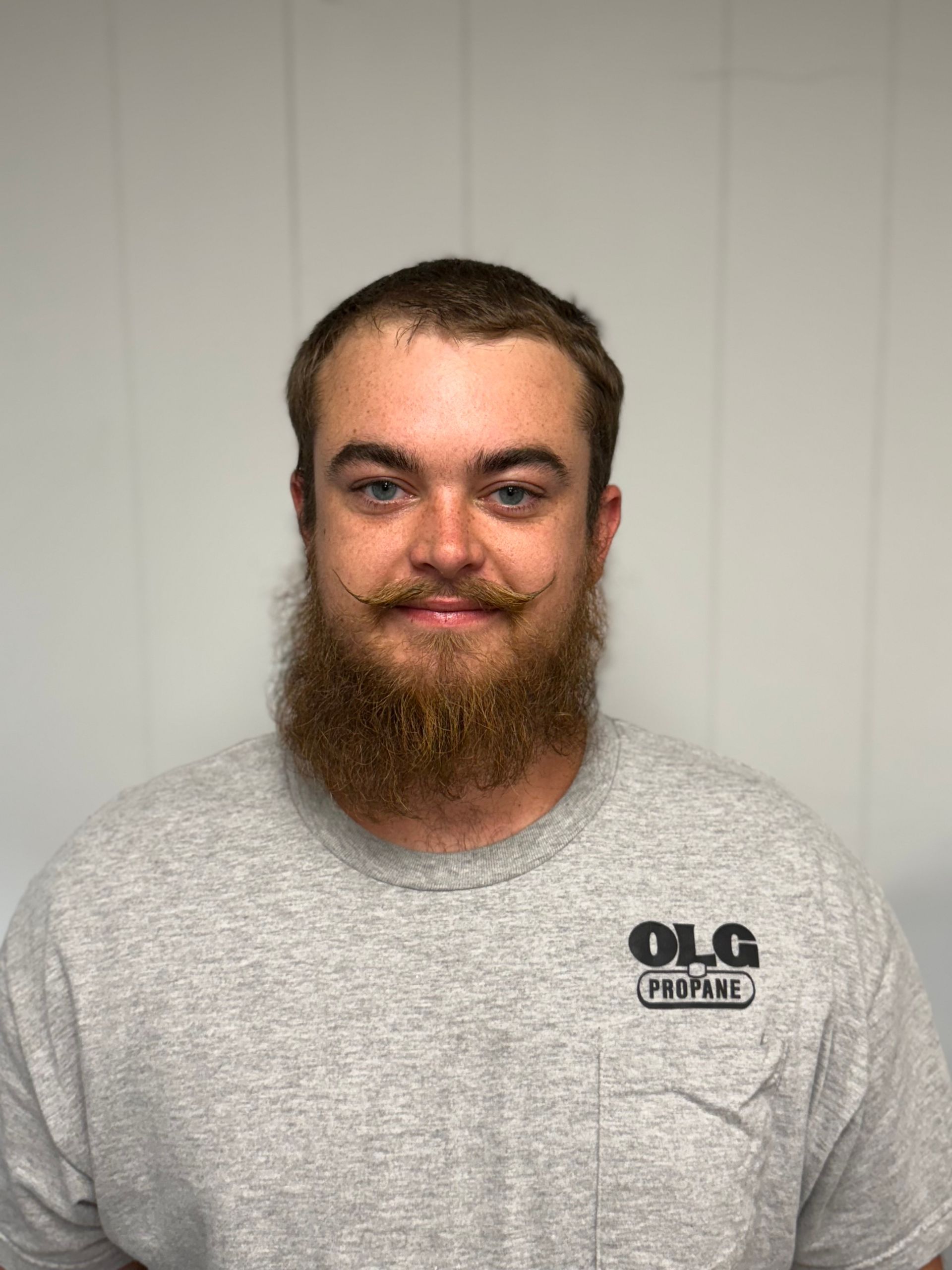 Man with brown hair, mustache, and beard wearing a gray shirt with a logo, smiling in front of a white wall.