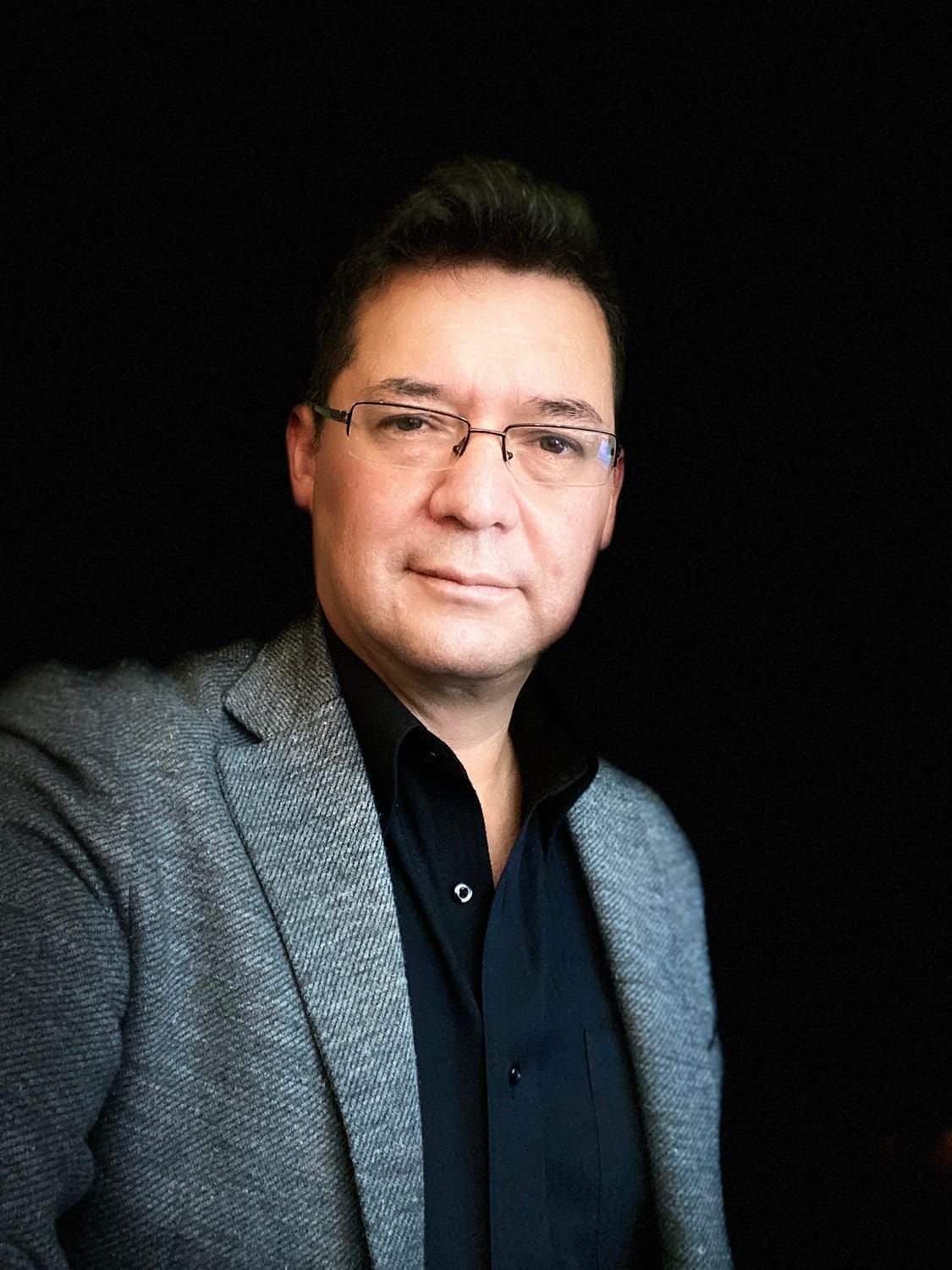 A man wearing glasses and a suit jacket is standing in front of a black background.