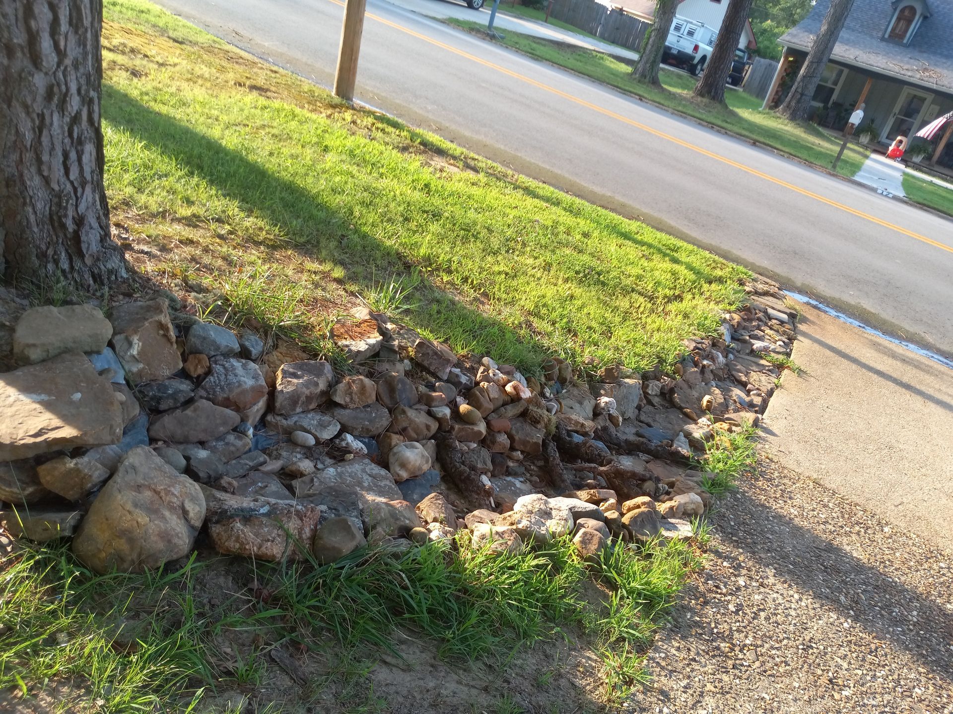 Rocks form a retaining wall along a grassy slope beside a street, with a tree on the left