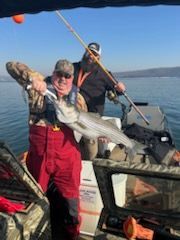 Two people on a boat, one smiling and holding up a large striped bass, the other standing behind holding a fishing rod.