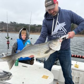 An adult and child on a boat hold up a large striped bass fish caught on a lake.