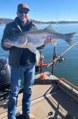 A person on a boat holding a large striped bass with a lake and clear sky in the background.