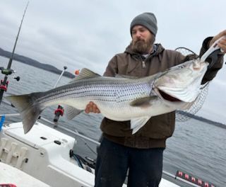 A person in a brown jacket and grey beanie holds a large, striped bass on a boat on a cloudy day.