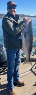 A person wearing a jacket and hat smiles while holding a large, striped fish on a boat on a sunny day.