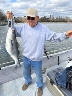 An individual on a boat holding up a large striped bass with both hands.